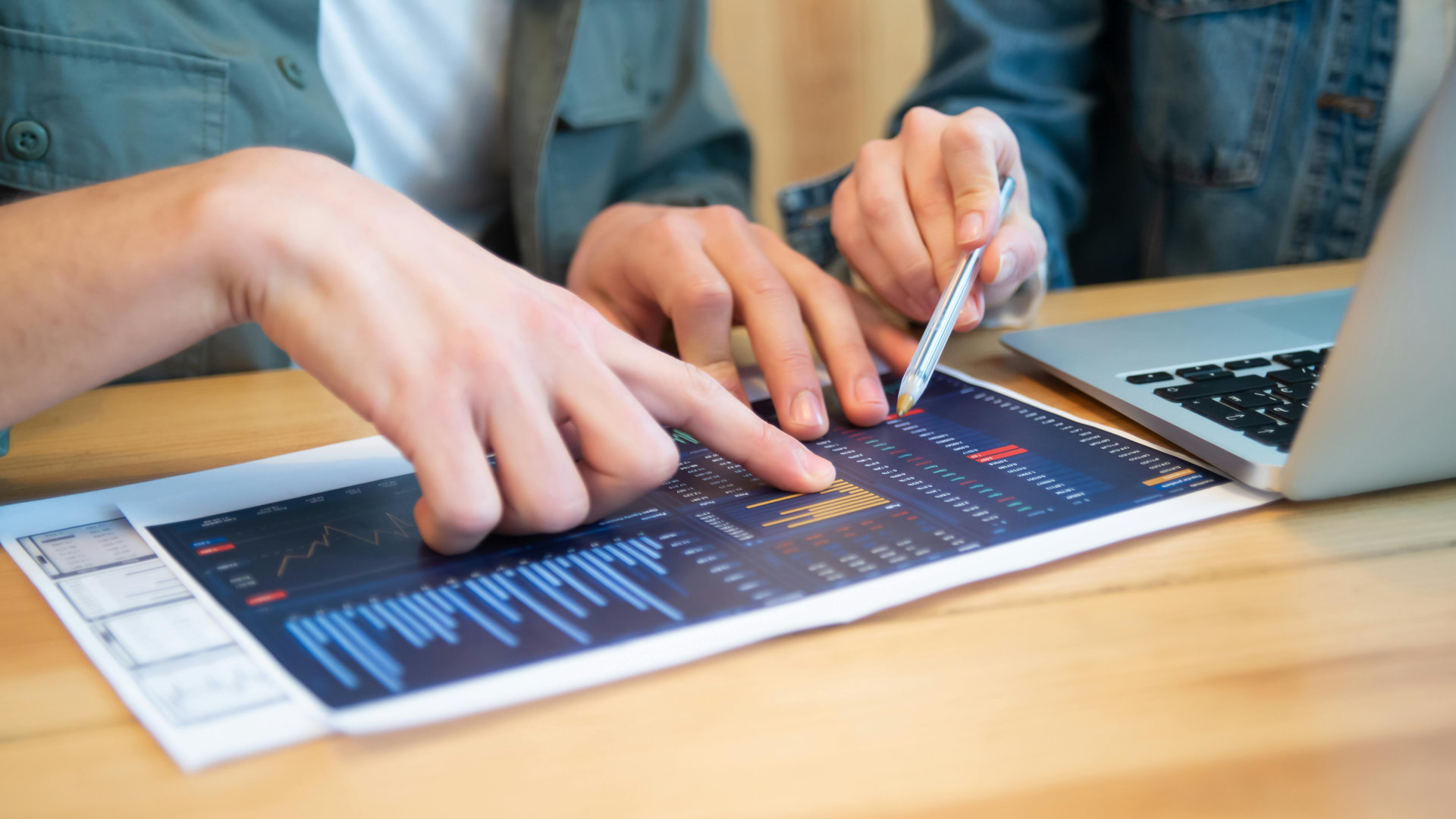 Two people reviewing a financial chart printed on paper, pointing and discussing data trends and bar graphs beside an open laptop on a wooden desk.