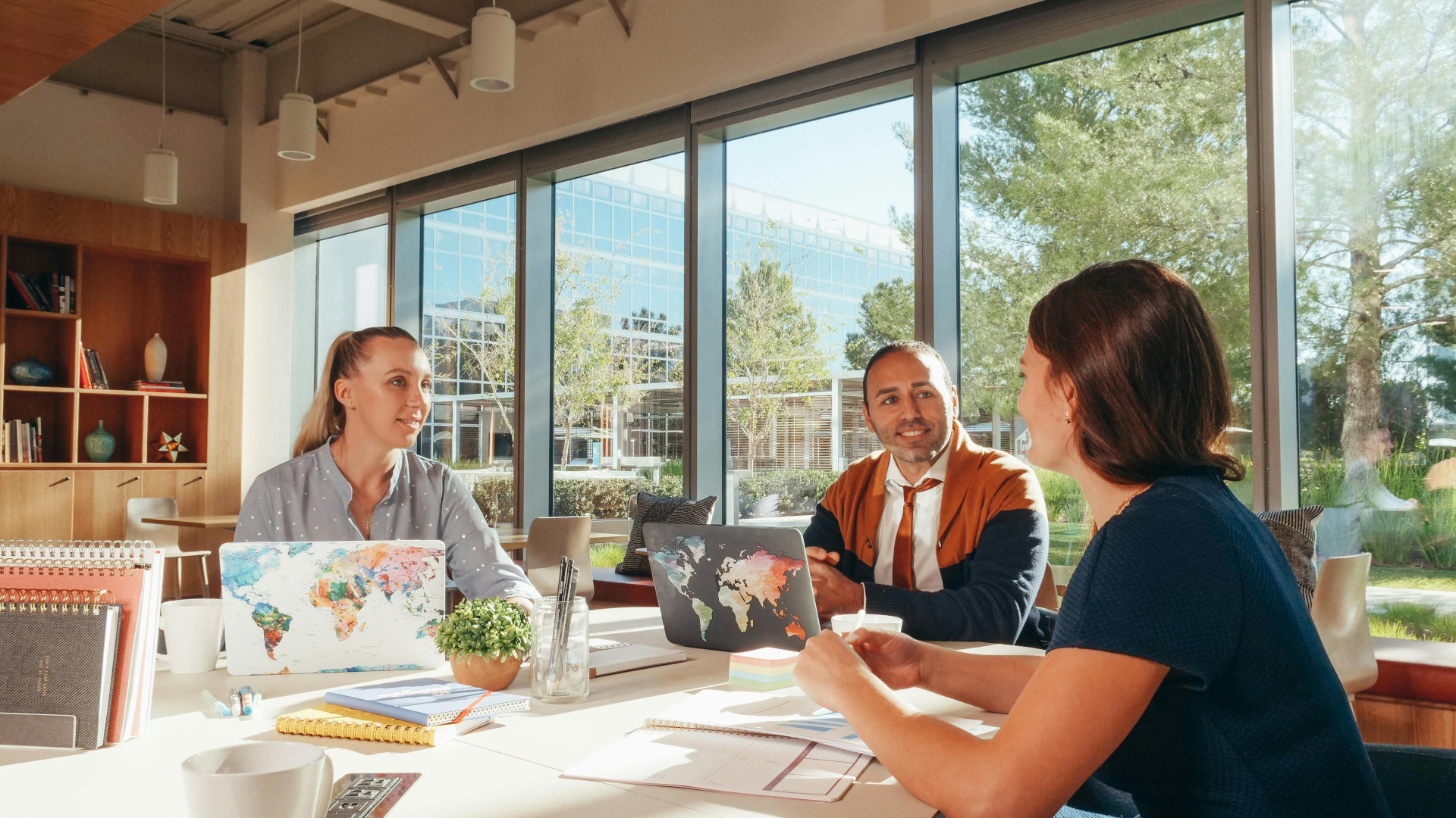 A small group of professionals meeting around a table with laptops in a bright modern office, collaborating in front of large windows overlooking trees and glass buildings.